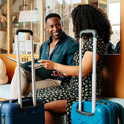 A man and a woman sitting together on a bench looking into a book with travel luggage next to them