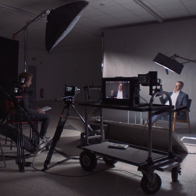 Ein Filmset mit Kameras, Beleuchtungsgeräten und einem Monitor, der einen sitzenden Interviewpartner in einer Studioumgebung zeigt.