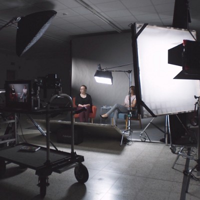 Ein Filmset mit zwei sitzenden Frauen in stilvollen Stühlen mit professioneller Beleuchtung und Ausrüstung vor einem grauen Hintergrund.