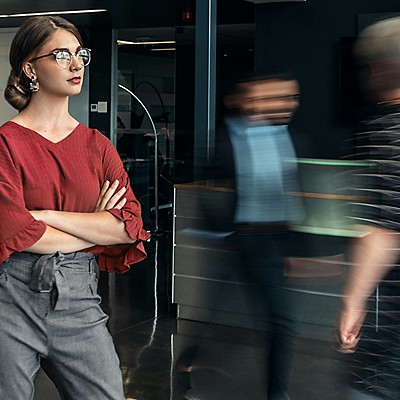 A woman wearing glasses stands in a modern office, surrounded by people